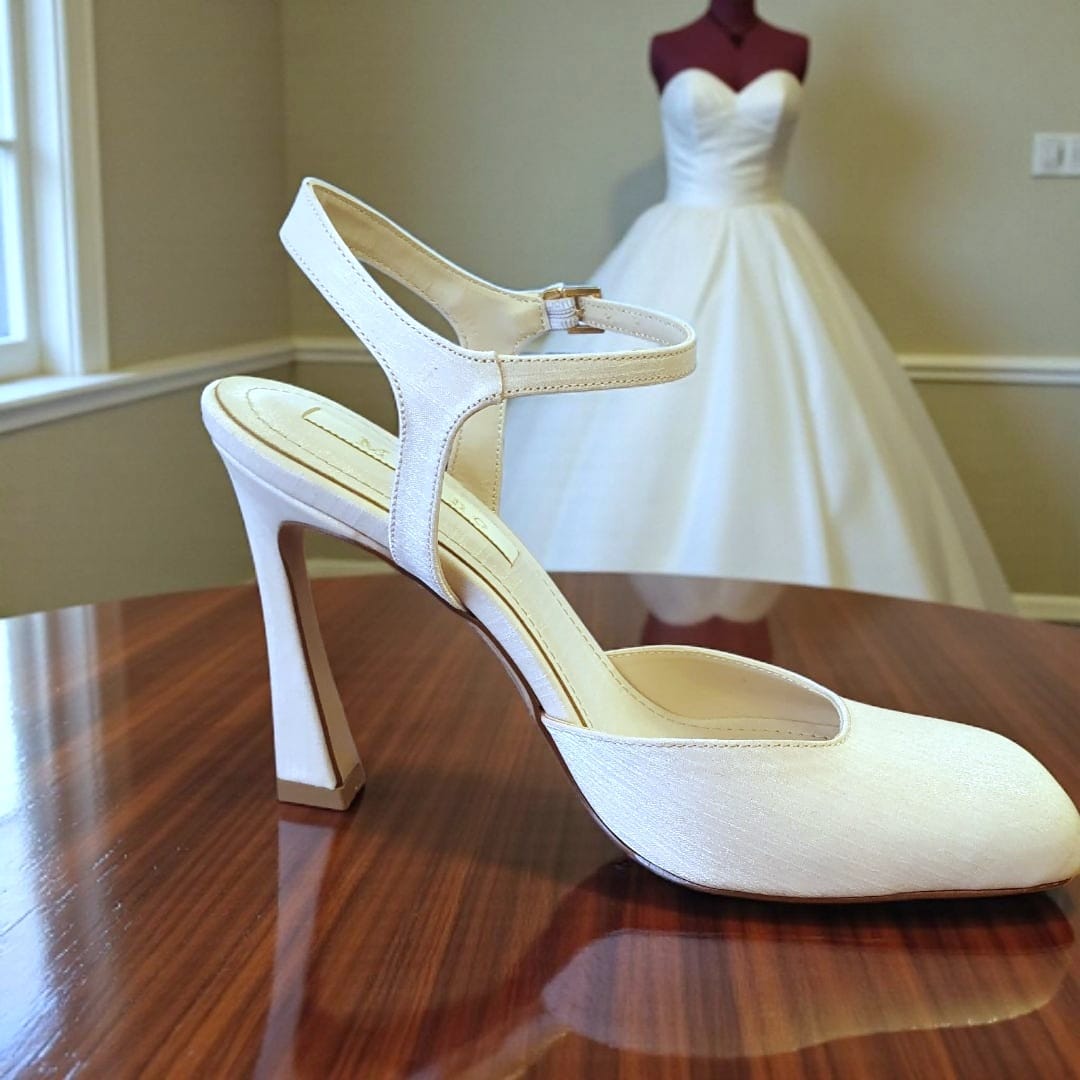 Pearl White high-heeled shoe on a wooden surface with a blurred wedding dress in the background.