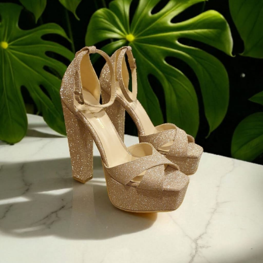 Gold glitter high-heeled shoes with bow detail on a marble surface with green leaves in the background