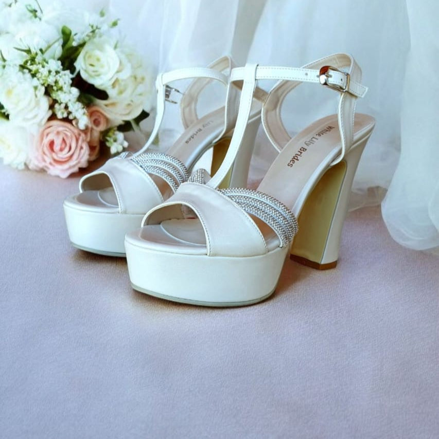 White wedding shoes with platform and high heel on a light pink surface with white flowers.