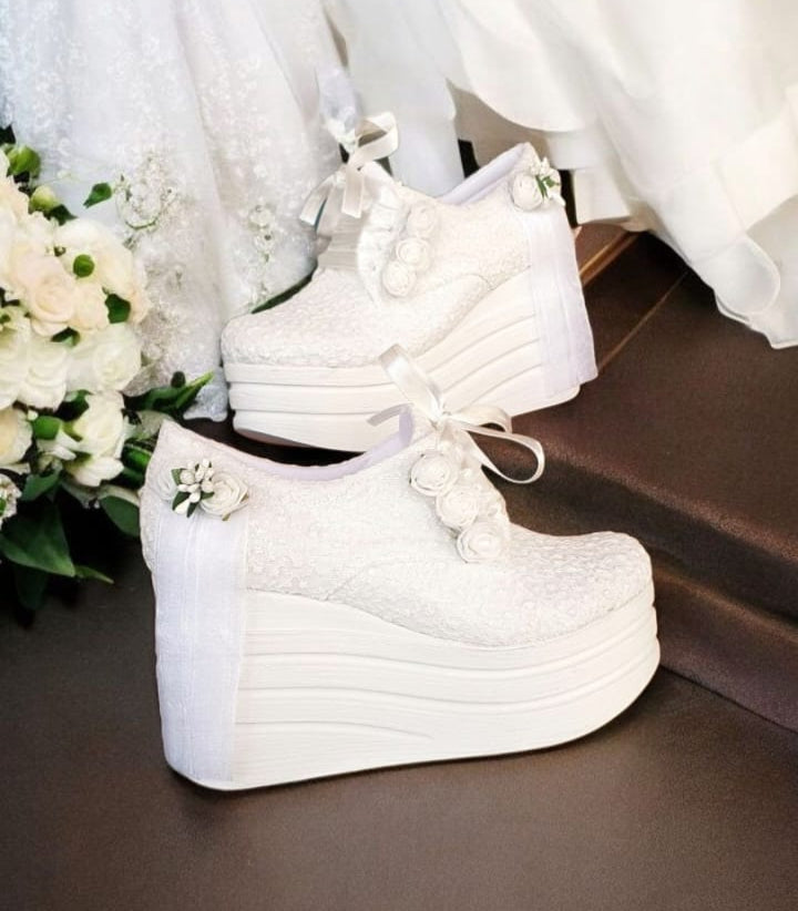 White wedding shoes with decorative ribbons next to a bouquet on a dark surface.