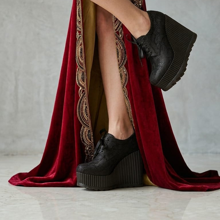 A model wearing a floor-length red velvet gown with gold embroidery and a high slit, paired with black platform lace-up shoes, posed against a textured gray studio wall.