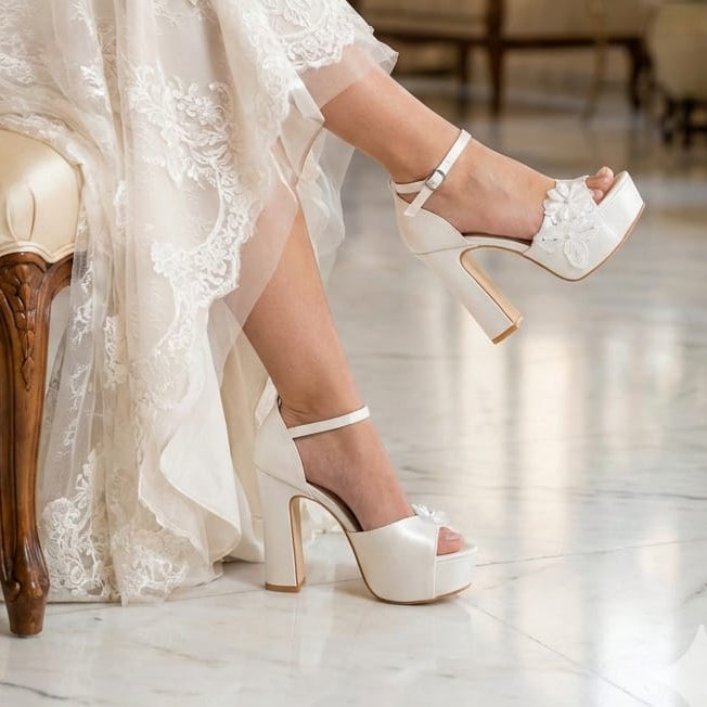 A bride with crossed legs wearing luxury white platform block heels featuring floral embellishments from the Wedding and Bridal Shoes Collection by White Lily Brides, seated on a white marble floor
