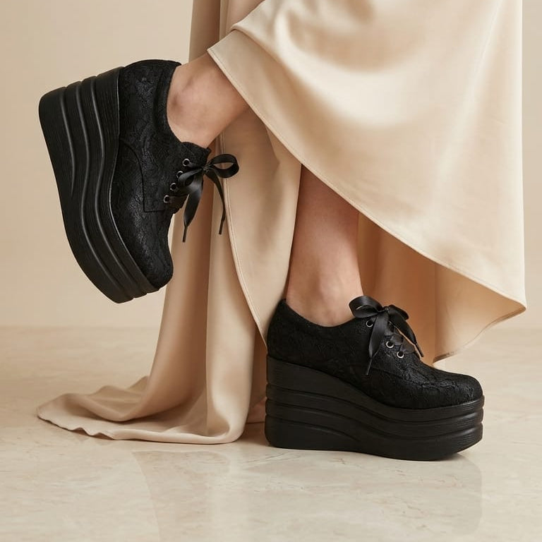 A person wearing black lace-up platform sneakers with textured uppers and chunky black platform soles, styled with a flowing beige satin skirt against a neutral beige studio background.