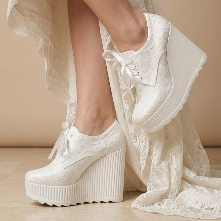 White lace-up platform wedge shoes with textured fabric and thick ribbed soles, shown on a model wearing a white lace dress against a beige studio background.