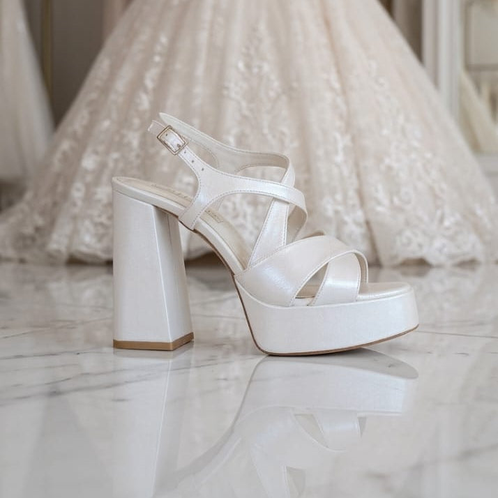 A shimmering pearl white bridal platform shoe with a 12cm block heel and elegant crisscross straps, shown reflecting on a polished marble floor. In the soft-focus background, a detailed lace wedding gown stands on a mannequin in a Beirut, Lebanon bridal boutique. Designed by White Lily Brides.