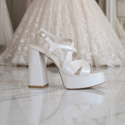 A shimmering pearl white bridal platform shoe with a 12cm block heel and elegant crisscross straps, shown reflecting on a polished marble floor. In the soft-focus background, a detailed lace wedding gown stands on a mannequin in a Beirut, Lebanon bridal boutique. Designed by White Lily Brides.