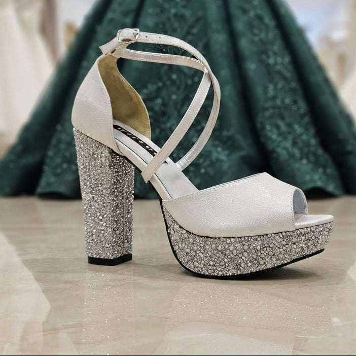 A Silver peep-toe high-heeled shoe with a sparkling silver rhinestone platform and block heel, displayed in a bridal shop with a green gown blurred in the background. Lebanon