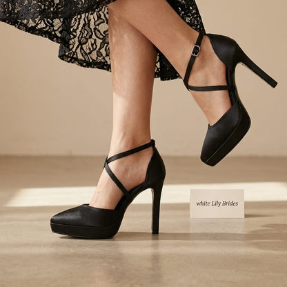 Black platform high-heel pumps with ankle straps worn by a model in a black lace dress, shown against a warm beige studio background with a small label reading 'white Lily Brides'.