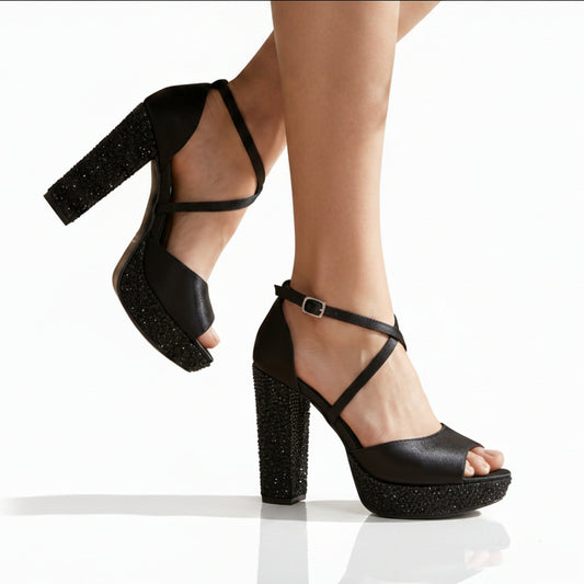 Black platform high-heel sandals with ankle straps and glittery block heels, shown on a model's feet against a warm beige studio background.