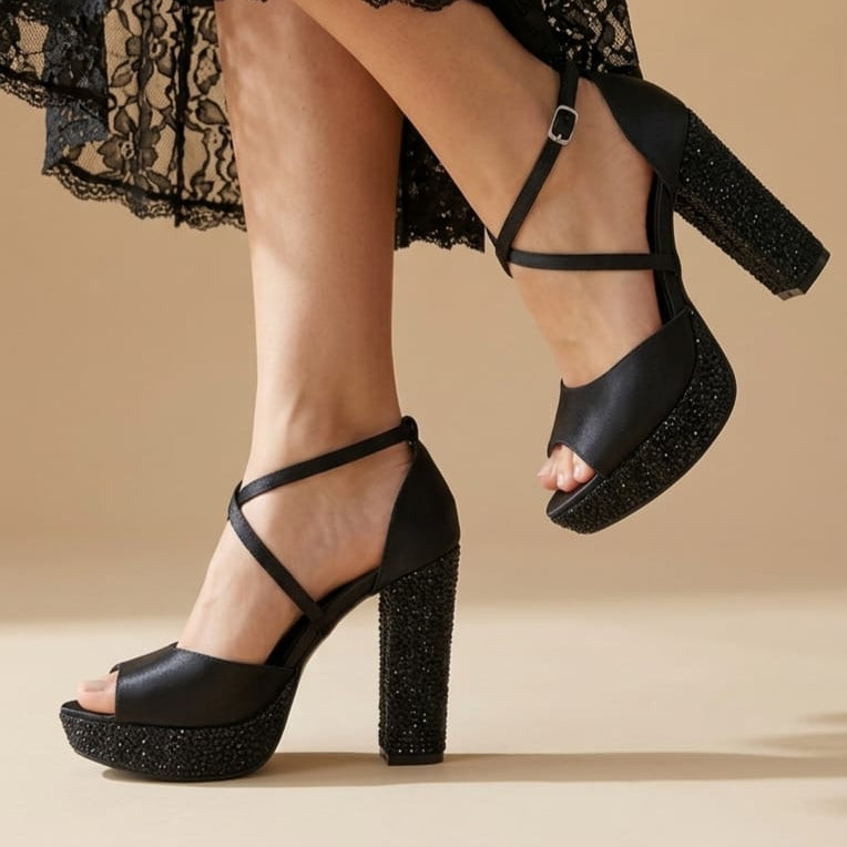Black strappy platform sandals with glittery block heels, shown on a model wearing a black lace skirt against a beige studio background.