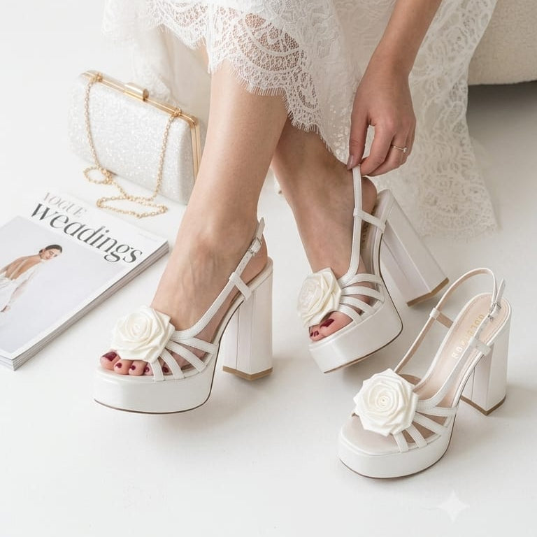 A woman in a white lace wedding dress sits on a white stool, wearing white Bridal platform high-heel sandals with large fabric flowers on the toes. She is holding one of the sandals, and another is placed beside her on the floor next to a magazine and a small handbag.