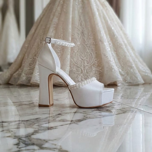A White Lily Brides white bridal shoe with pearl-embellished straps, a high block heel, and a platform, resting on a glossy, reflective marble floor. A softly blurred, luxurious lace wedding gown 