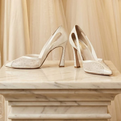 Pair of high-heeled wedding shoes on a marble pedestal with wedding dresses in the background