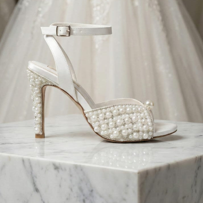 White pearl-studded high-heeled shoe on a marble surface with a wedding dress in the background.