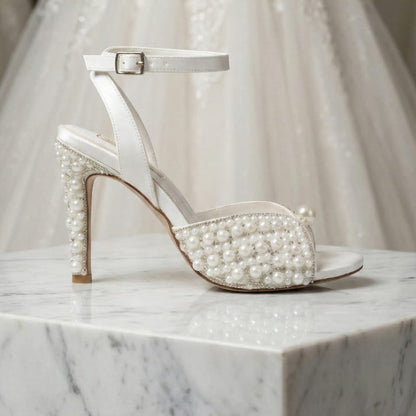 White pearl-studded high-heeled shoe on a marble surface with a wedding dress in the background.
