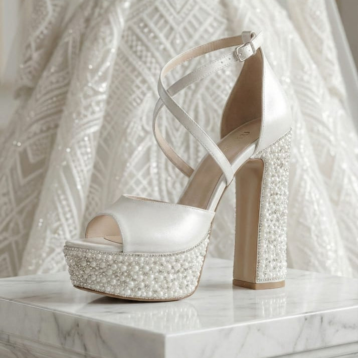 Pearl White high-heeled sandal with pearl embellishments on a marble surface, with a white wedding lace dress in the background. Beirut
