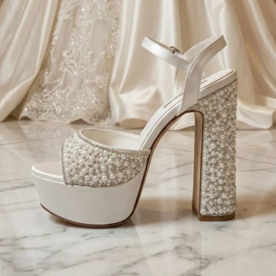 Pearl White high-heeled shoes with crystal embellishments in front of a white lace wedding dress.