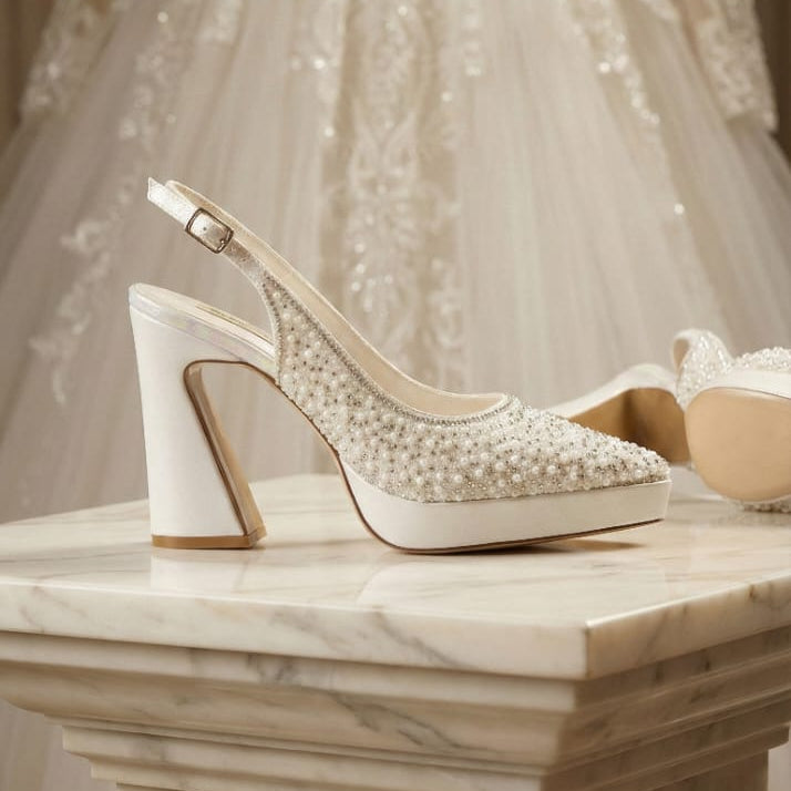 Pearl White high-heeled shoes with pearl embellishments on a marble surface, with a wedding dress in the background. Beirut