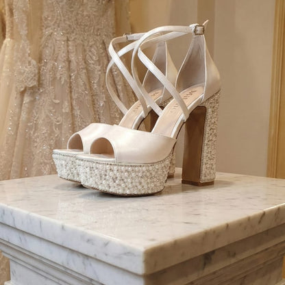 Pearl White high-heeled wedding shoes with embellishments on a marble surface, with a blurred background of a wedding dress.