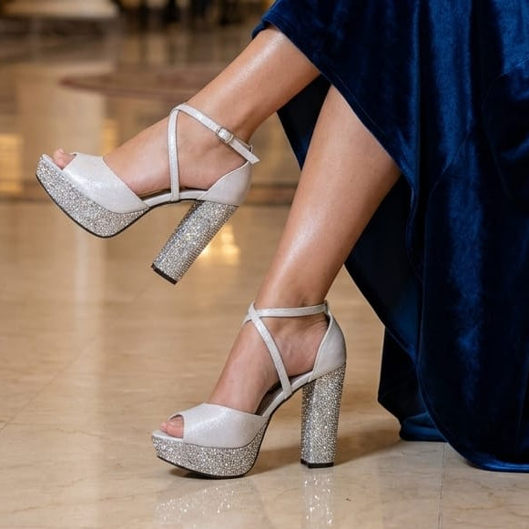 A person seated in a grand indoor venue wearing a flowing navy blue dress and Silver strappy high-heeled sandals with glittery block heels. The setting includes marble columns, a piano, and a large floral arrangement in the background.
