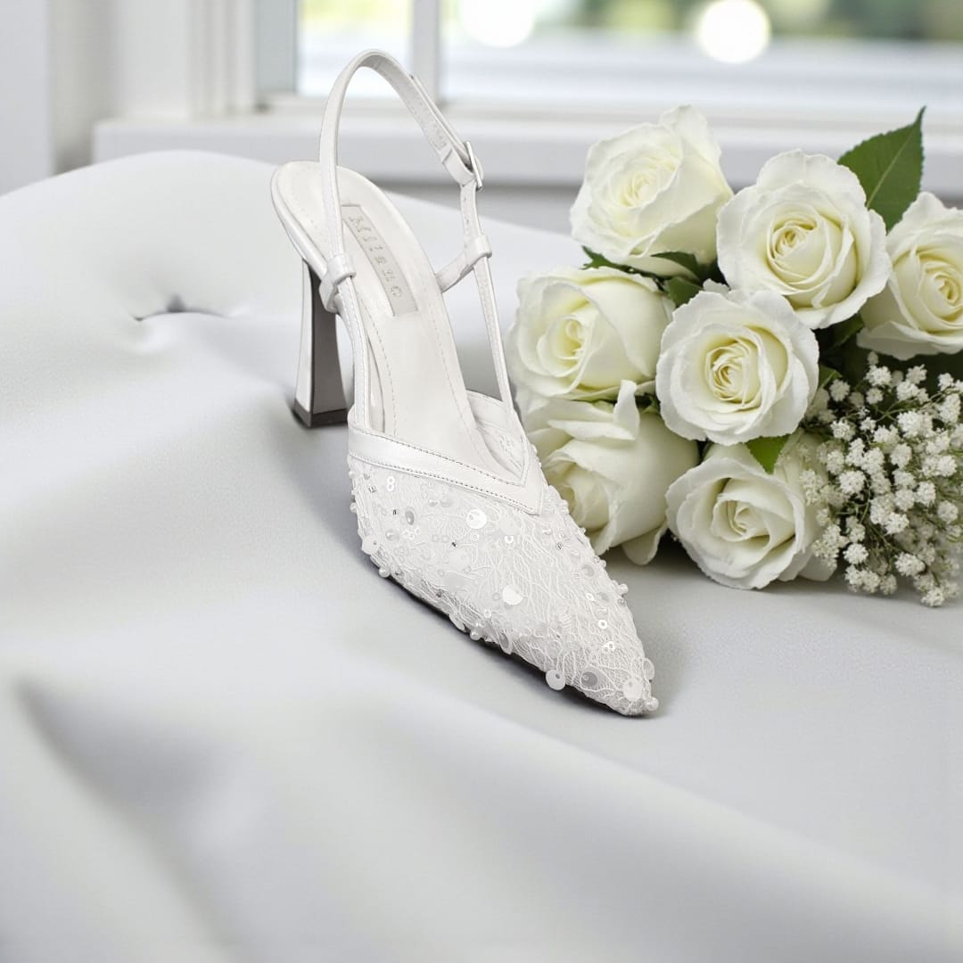 White wedding shoe with pearl details next to a bouquet of white roses on a light surface.