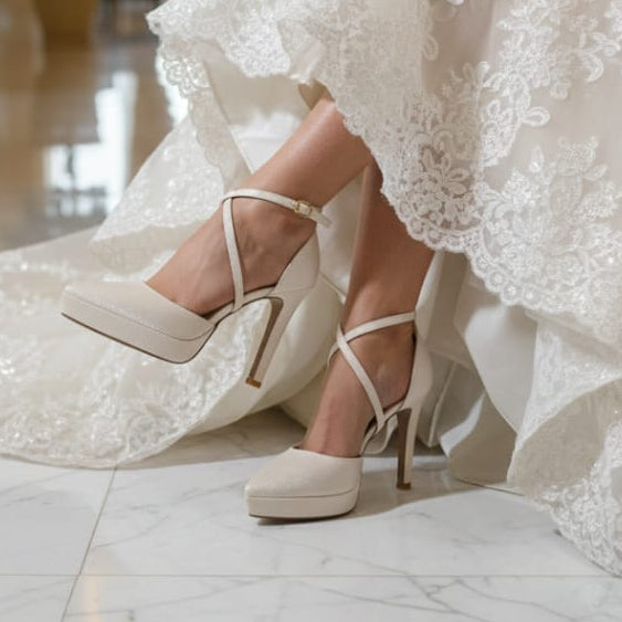Bride in a white lace dress sitting on a chair with white high-heeled shoes.