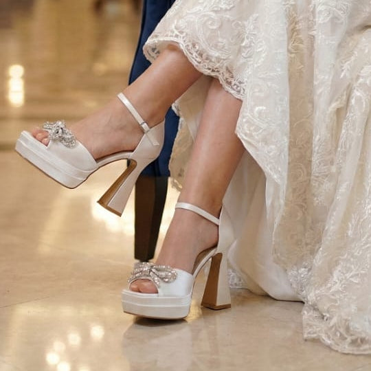 White high-heeled shoes with decorative straps worn by a person in a formal setting.