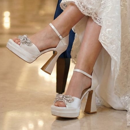 White high-heeled shoes with decorative straps worn by a person in a formal setting.
