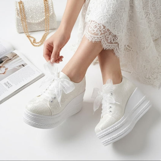 White sneakers with bows worn by a person in a white lace dress on a light background