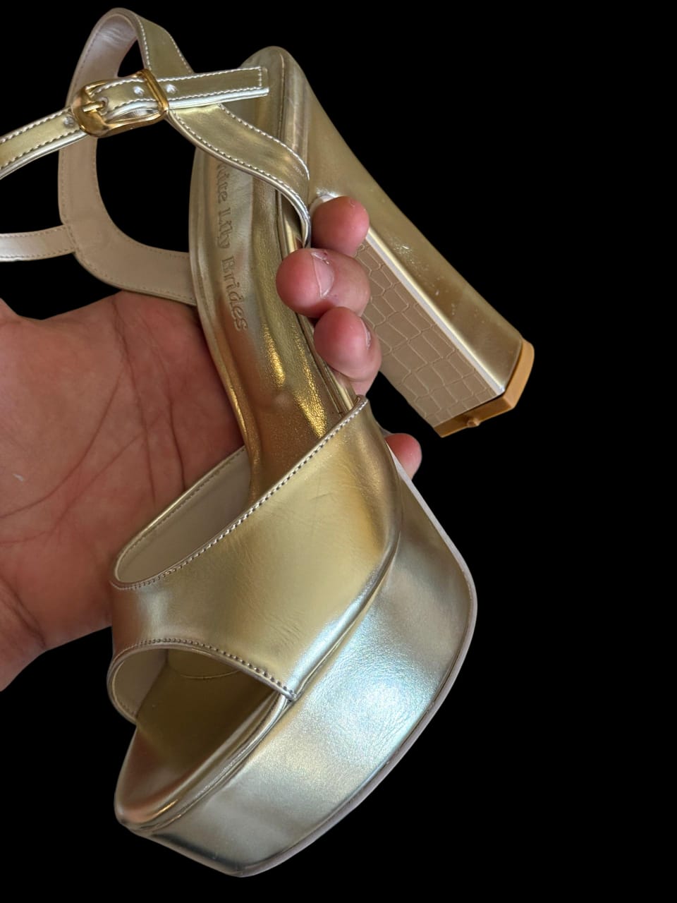 Gold high-heeled shoe held by a hand against a black background
