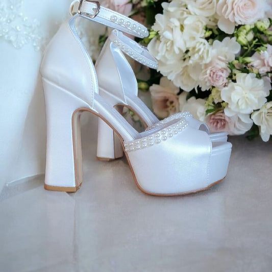 White wedding shoes with high heels and decorative straps next to a bouquet of flowers.