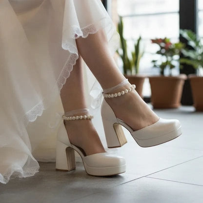 White wedding shoes with pearl straps worn by a person in a white dress.
