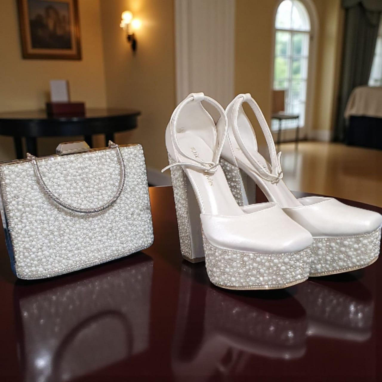 White high-heeled shoes with sparkling platform and matching handbag on a reflective surface.