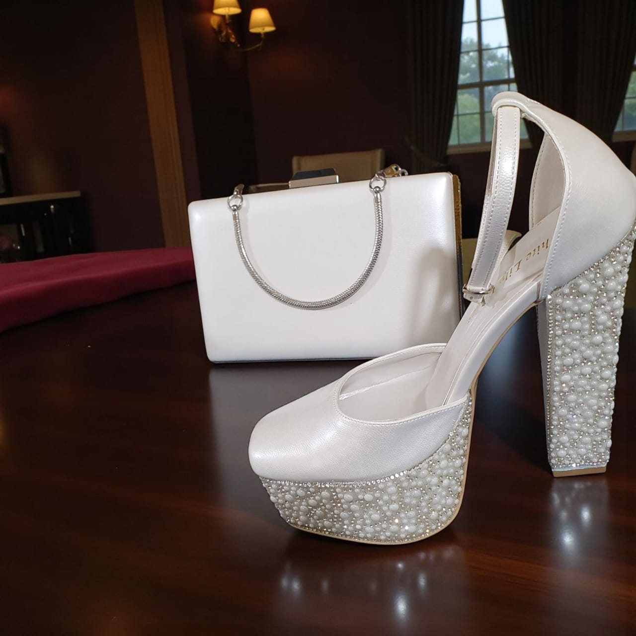 White high-heeled shoes with pearl embellishments and a matching handbag on a dark surface.