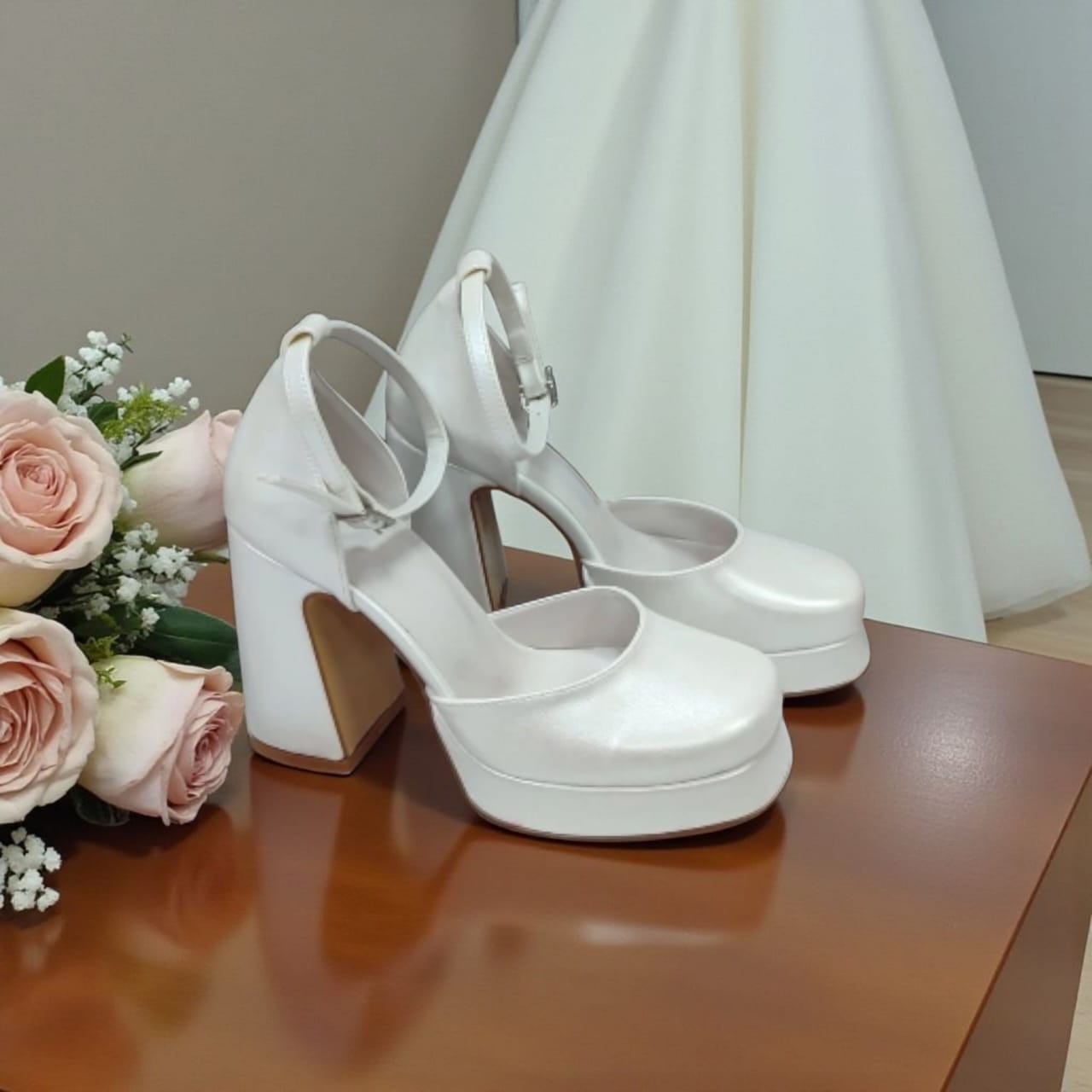 White wedding shoes with a bouquet of pink roses on a wooden surface.