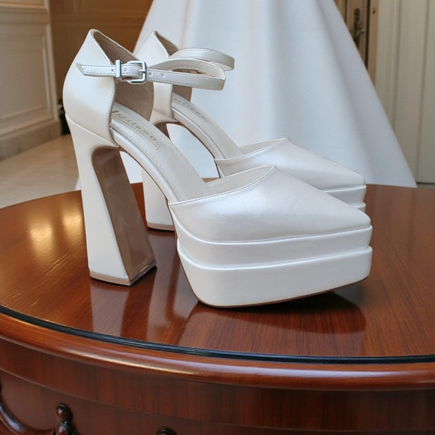 White wedding shoes with platform sole on a wooden table, white wedding dress in the background.