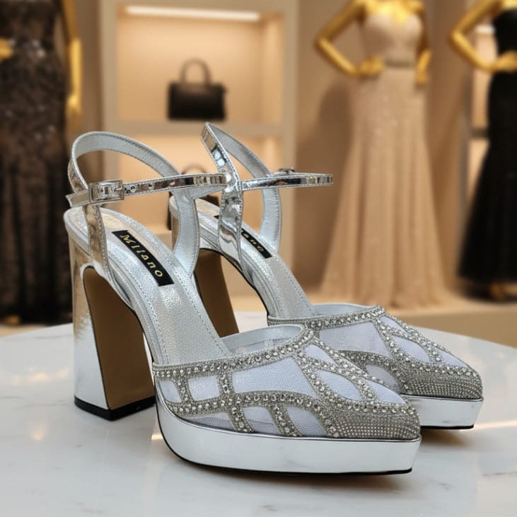 Silver high-heeled shoes with rhinestone details on a marble surface, with mannequins and bags in the background.