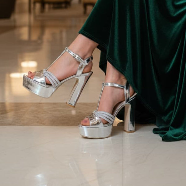 Person wearing a green dress and silver high heels in an elegant indoor setting with chandeliers.