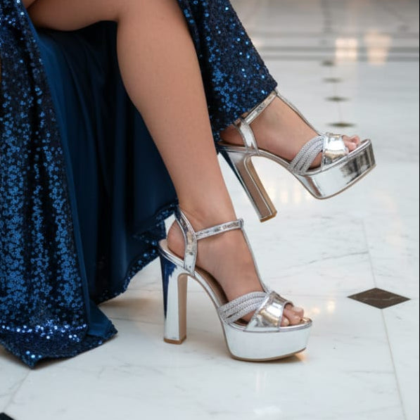 Silver high-heeled shoes worn with a blue sequin skirt on a light floor.