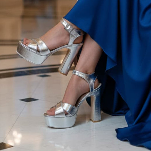Silver high-heeled platform shoes worn with a blue dress on a tiled floor.
