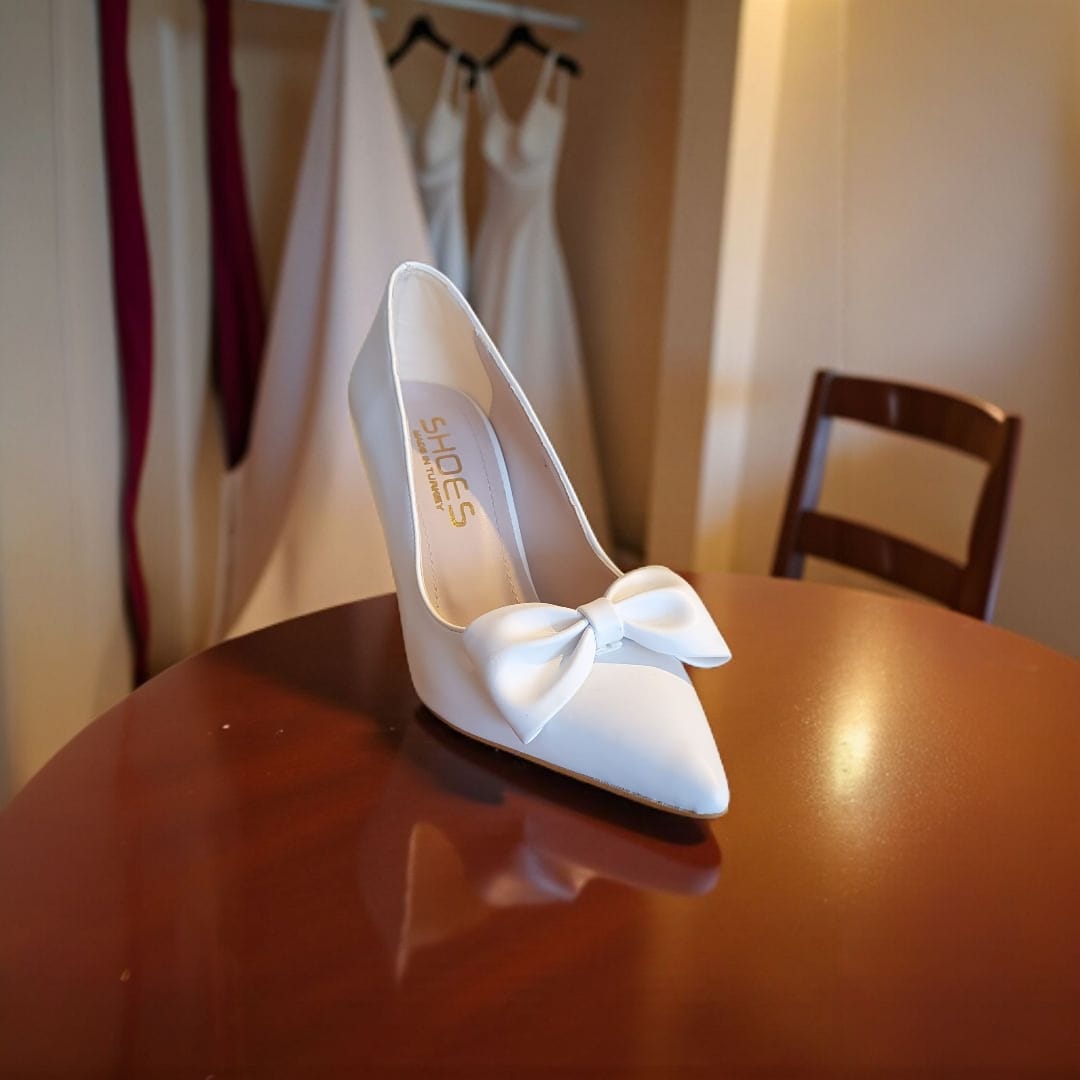 White high-heeled shoes with a bow on a wooden table, with wedding dresses in the background.
