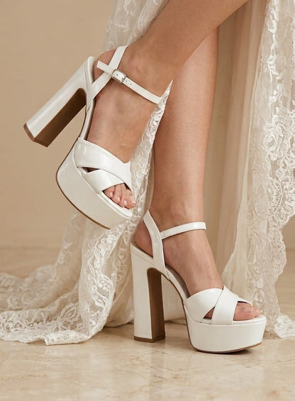 White platform block-heel sandals with ankle straps, shown on a model wearing a white lace dress in a warm indoor setting.