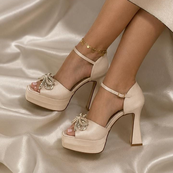 A pair of gold satin platform high-heeled sandals with ankle straps and jeweled toe embellishments, shown on a model's feet against a draped satin fabric background.