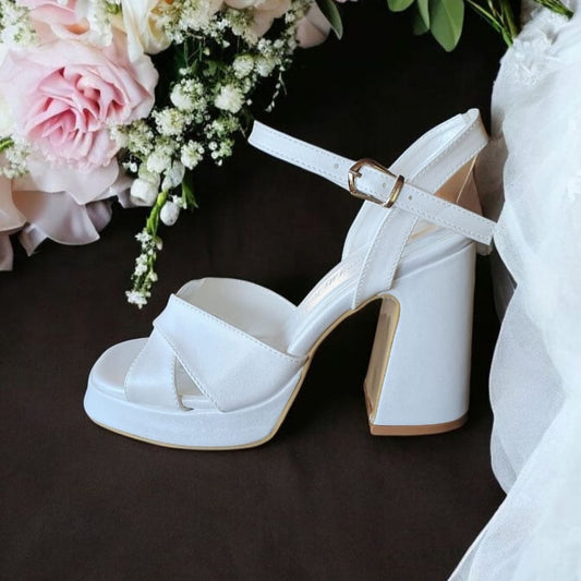 White block-heel bridal sandals with criss-cross straps and ankle buckle displayed on a dark tray.