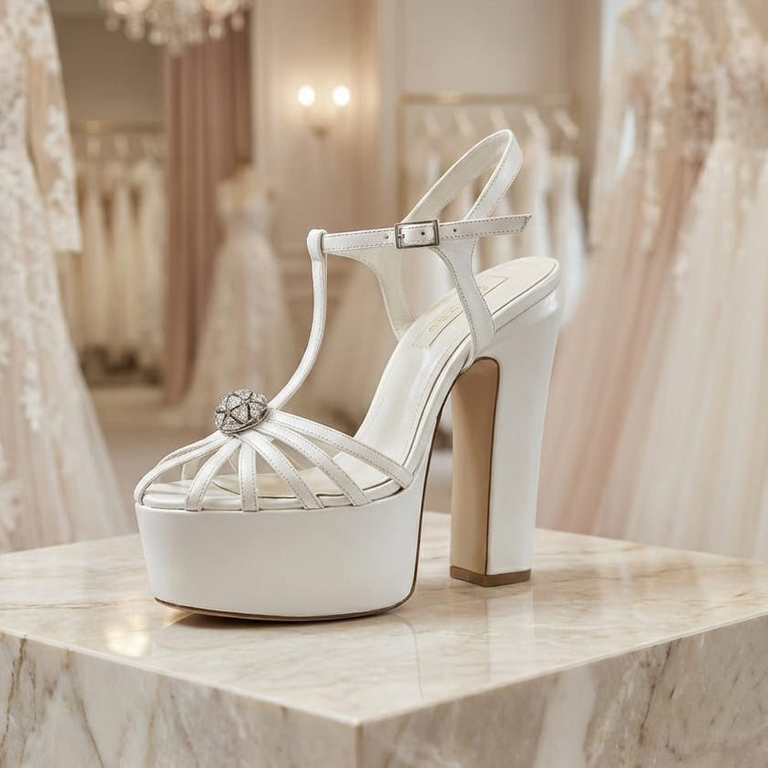White high-heeled shoe with a platform on a marble surface, with wedding dresses in the background.