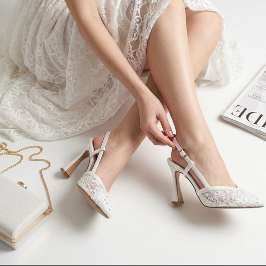 White lace high-heeled bridal shoes worn by a person sitting on a white surface with a magazine and jewelry box in the background.