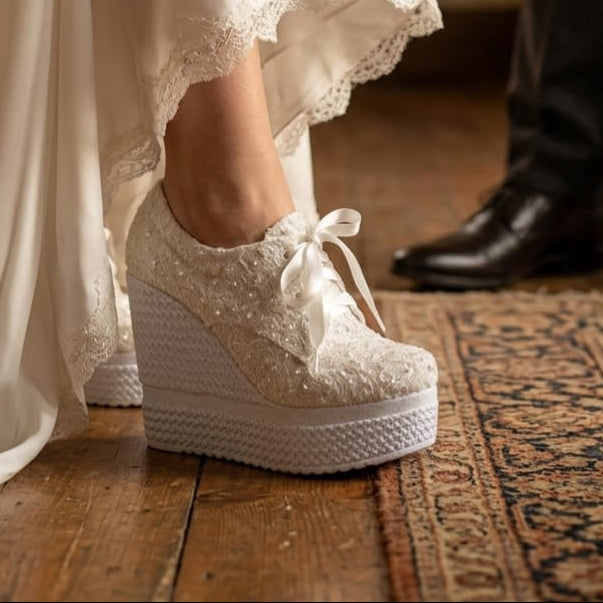 White lace wedding shoes worn with a flowing white dress on a wooden floor.