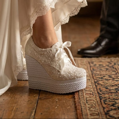 White lace wedding shoes worn with a flowing white dress on a wooden floor.