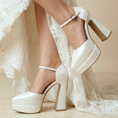 White platform high-heel shoes with ankle straps, shown on a model wearing a white lace wedding dress against a warm beige background.