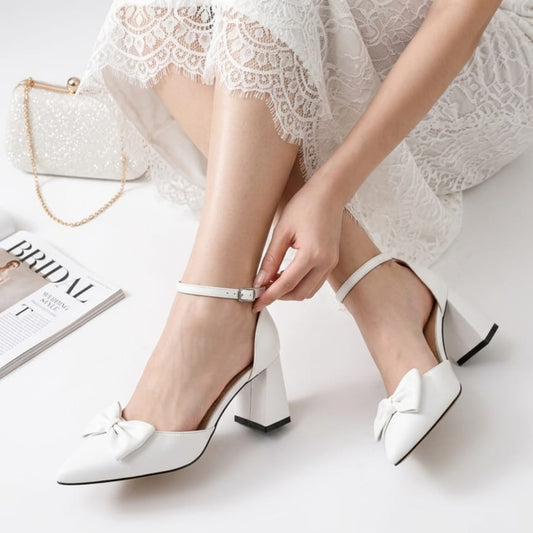 White wedding shoes with a bow being worn by a person, with a magazine and handbag in the background.